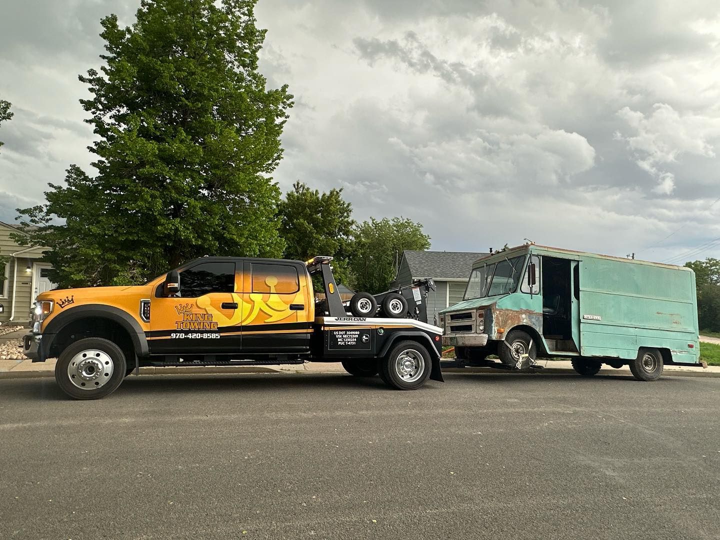 Yellow heavy-duty truck towing a dented mint-green box truck on a roadside under cloudy skies