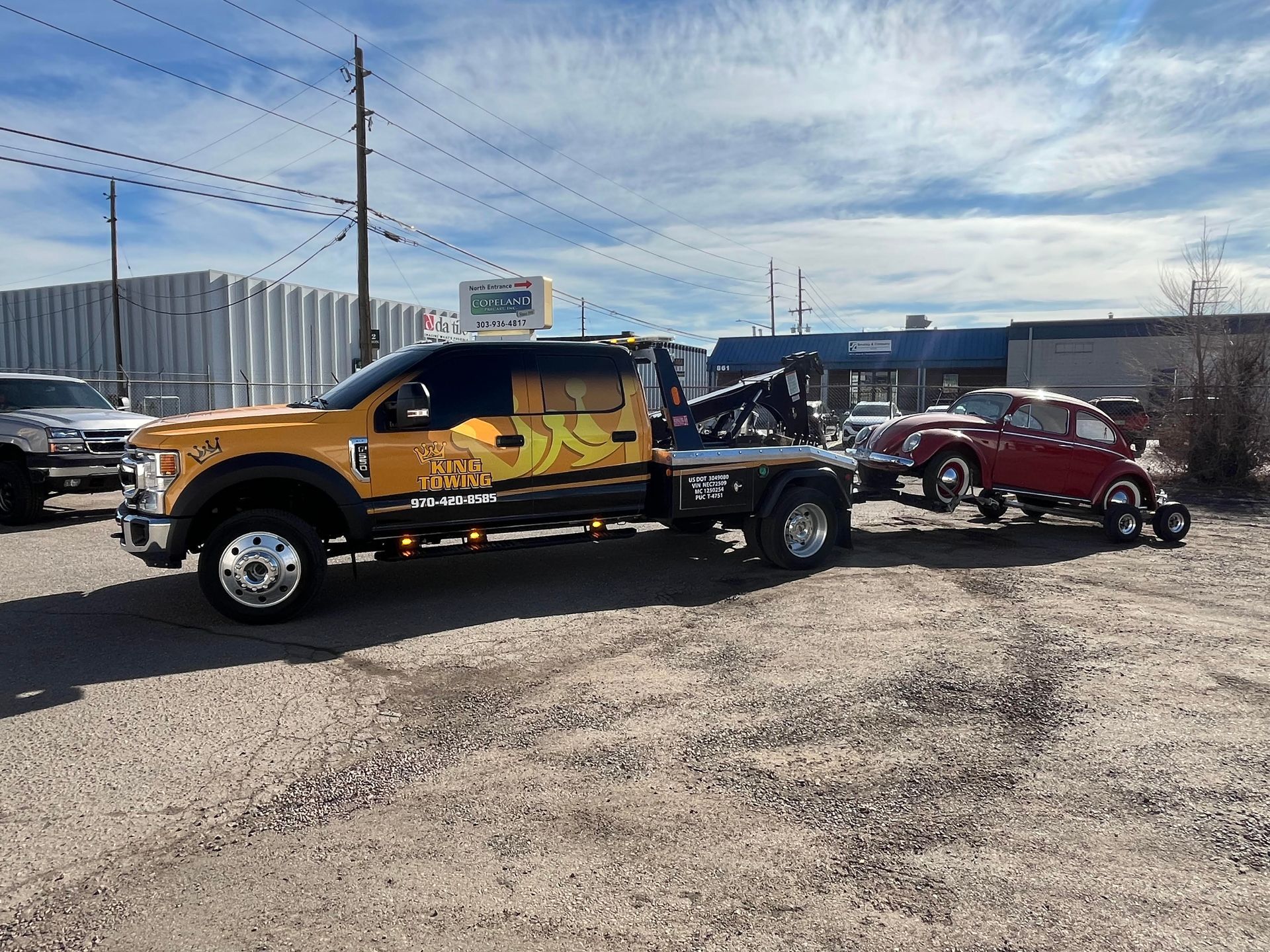 Yellow tow truck hauling a red car on a trailer in a gravel lot under a blue sky