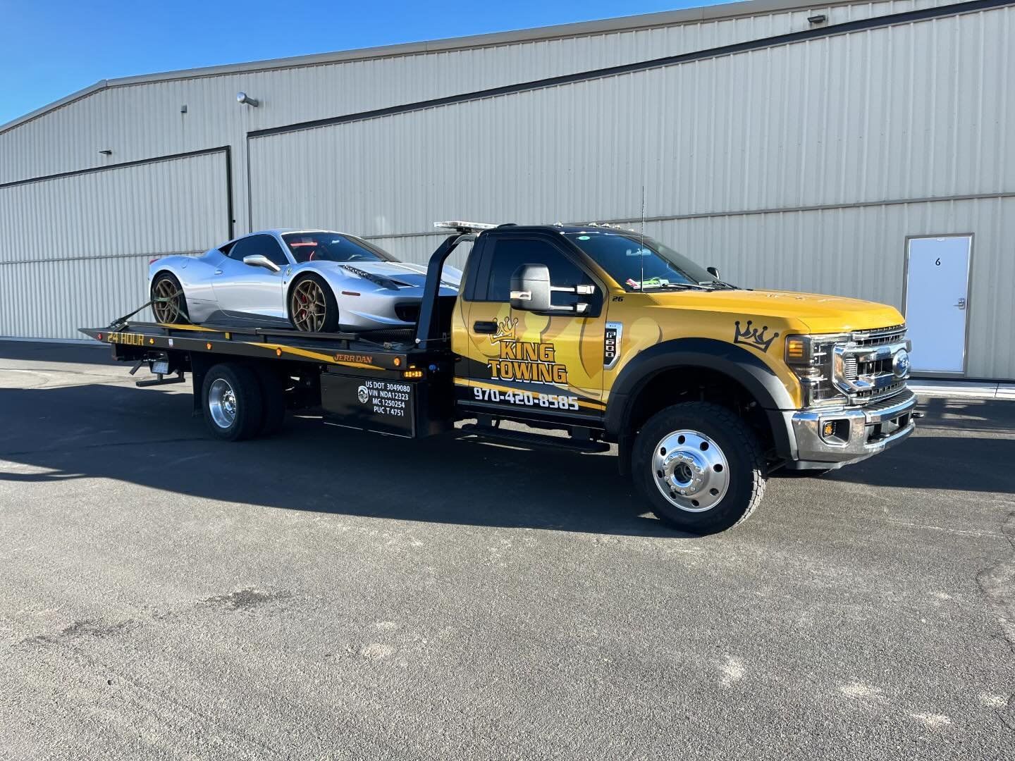 Yellow flatbed tow truck carrying a silver sports car outside a warehouse