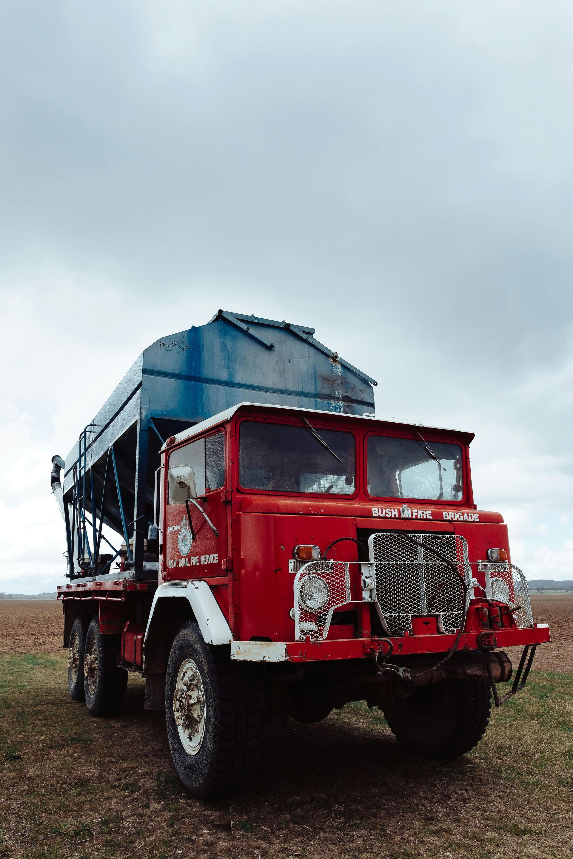 Red vintage truck with a blue cargo bed parked on a dirt field under cloudy sky