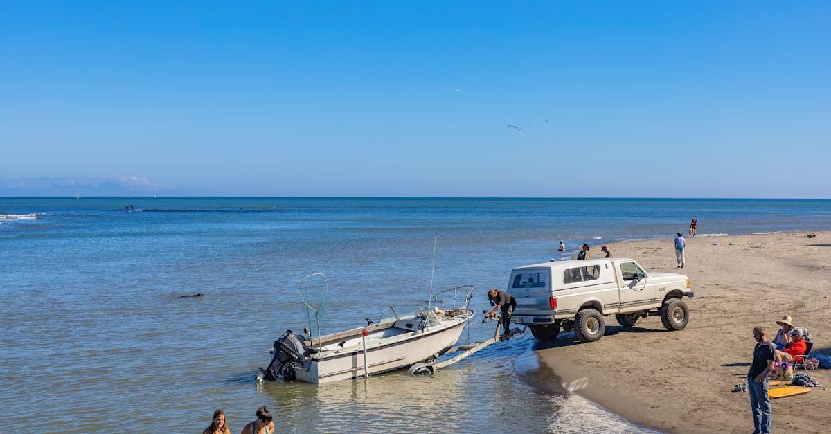 White SUV towing a small boat from a sandy shore into calm blue water, with people nearby