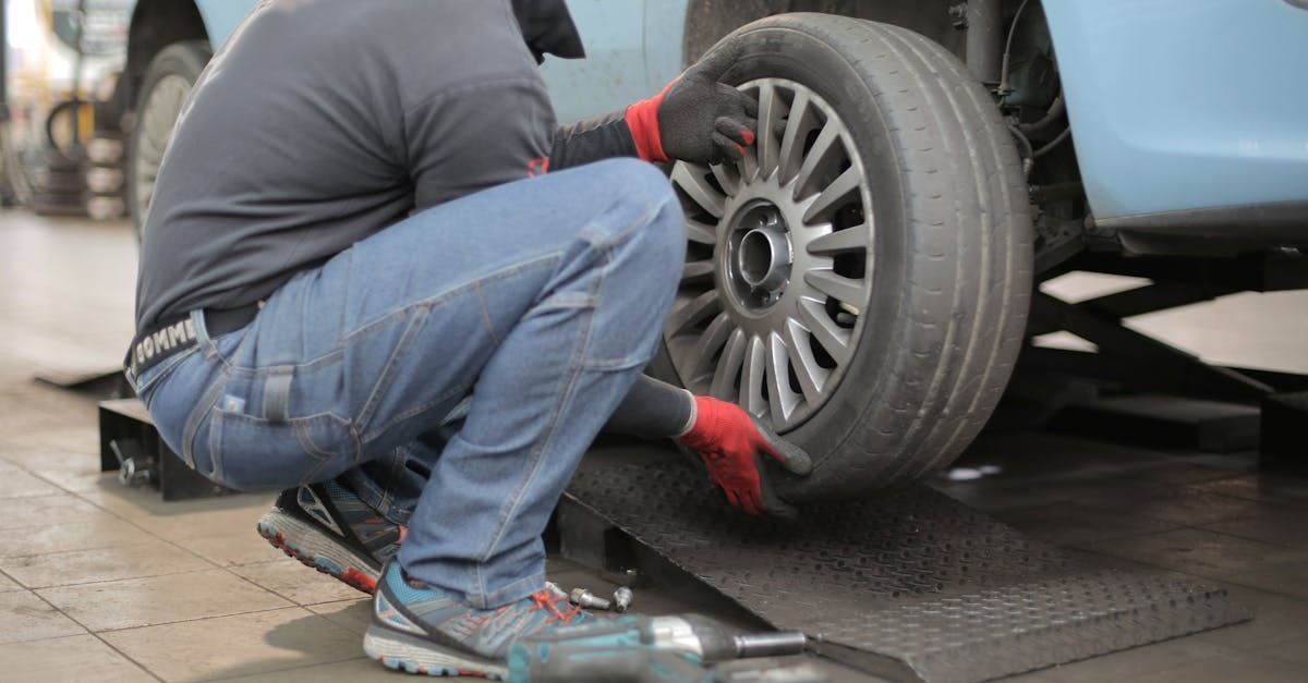 Mechanic crouches beside a car, inspecting a front wheel on a wet garage floor.