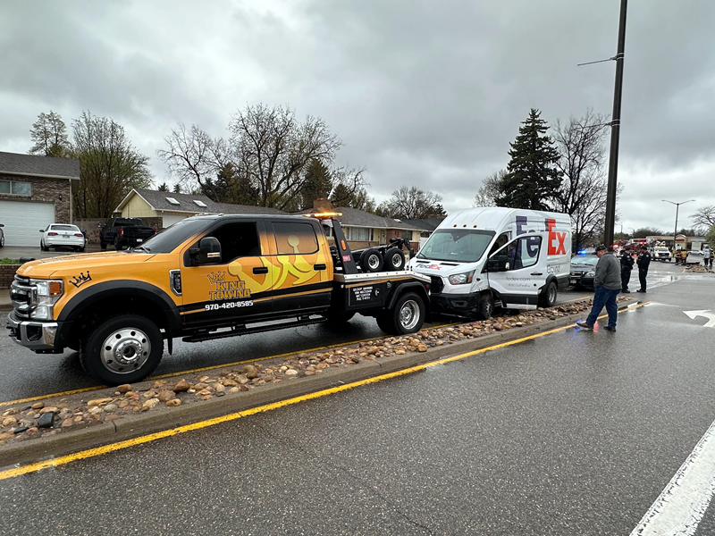 Tow truck towing a white van on a wet street after a crash, with workers nearby and cloudy skies