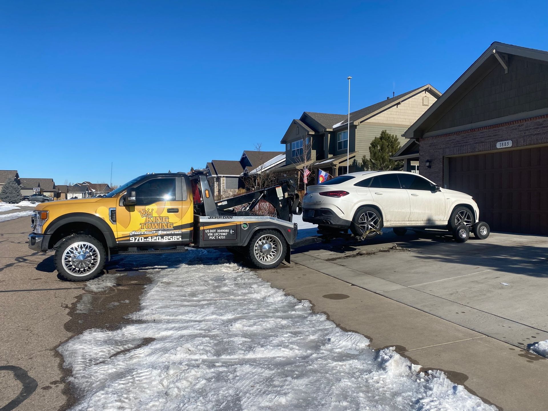 Yellow tow truck loading a white SUV in a residential driveway on a sunny day
