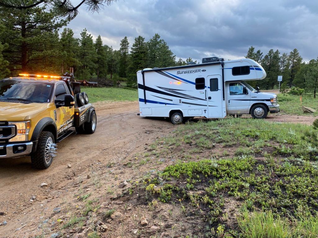 Yellow off-road truck and white camper truck parked on a muddy trail in a wooded area.