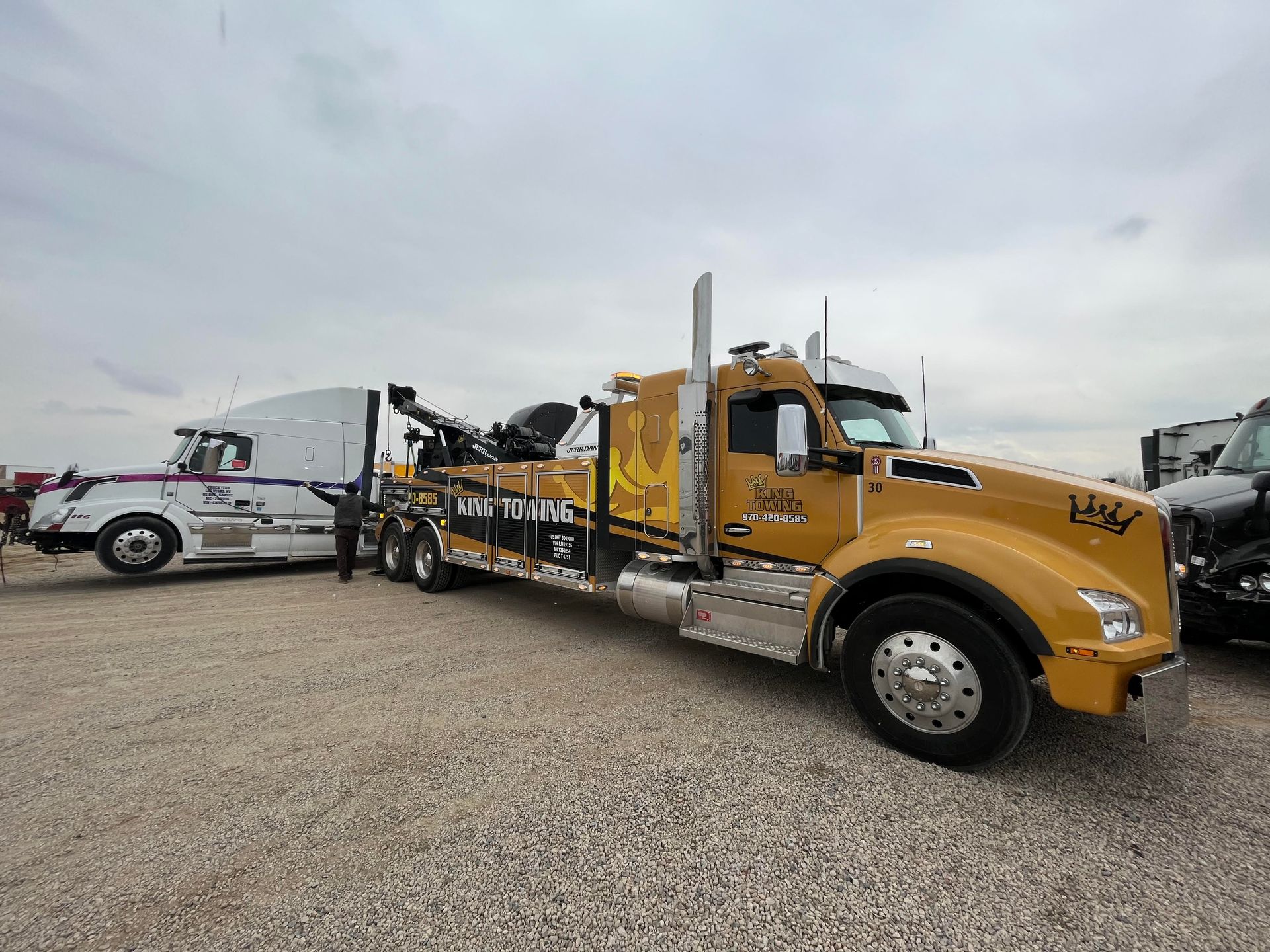 Yellow tow truck hauling a white truck on a gravel lot under cloudy skies