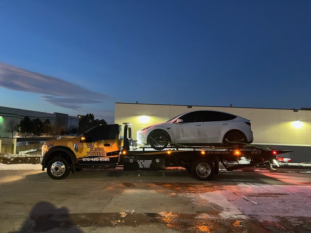 White SUV on a flatbed tow truck at dusk, parked outside a building with flashing lights.
