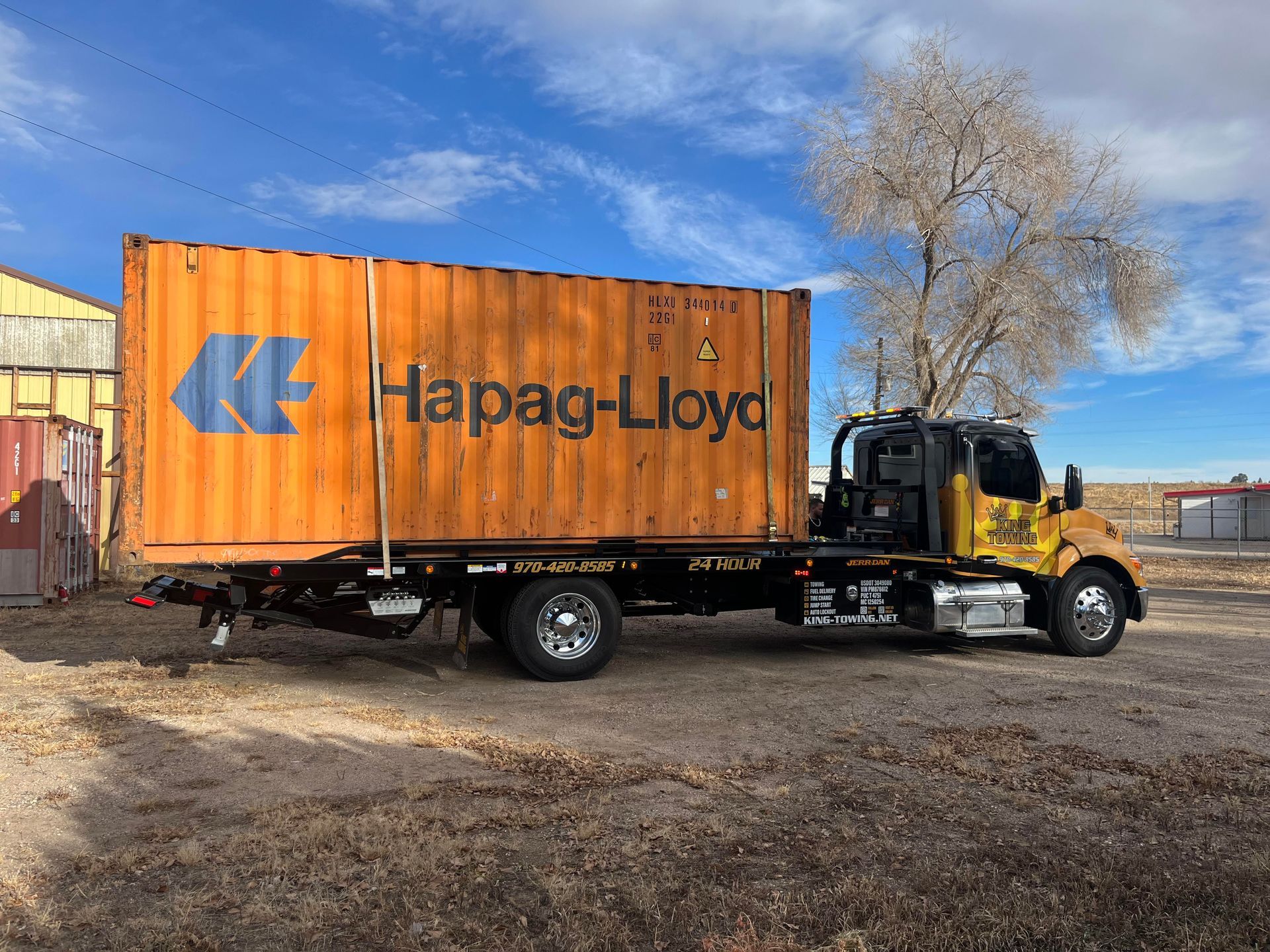 Black truck carrying an orange Hapag-Lloyd shipping container on a gravel lot under a blue sky