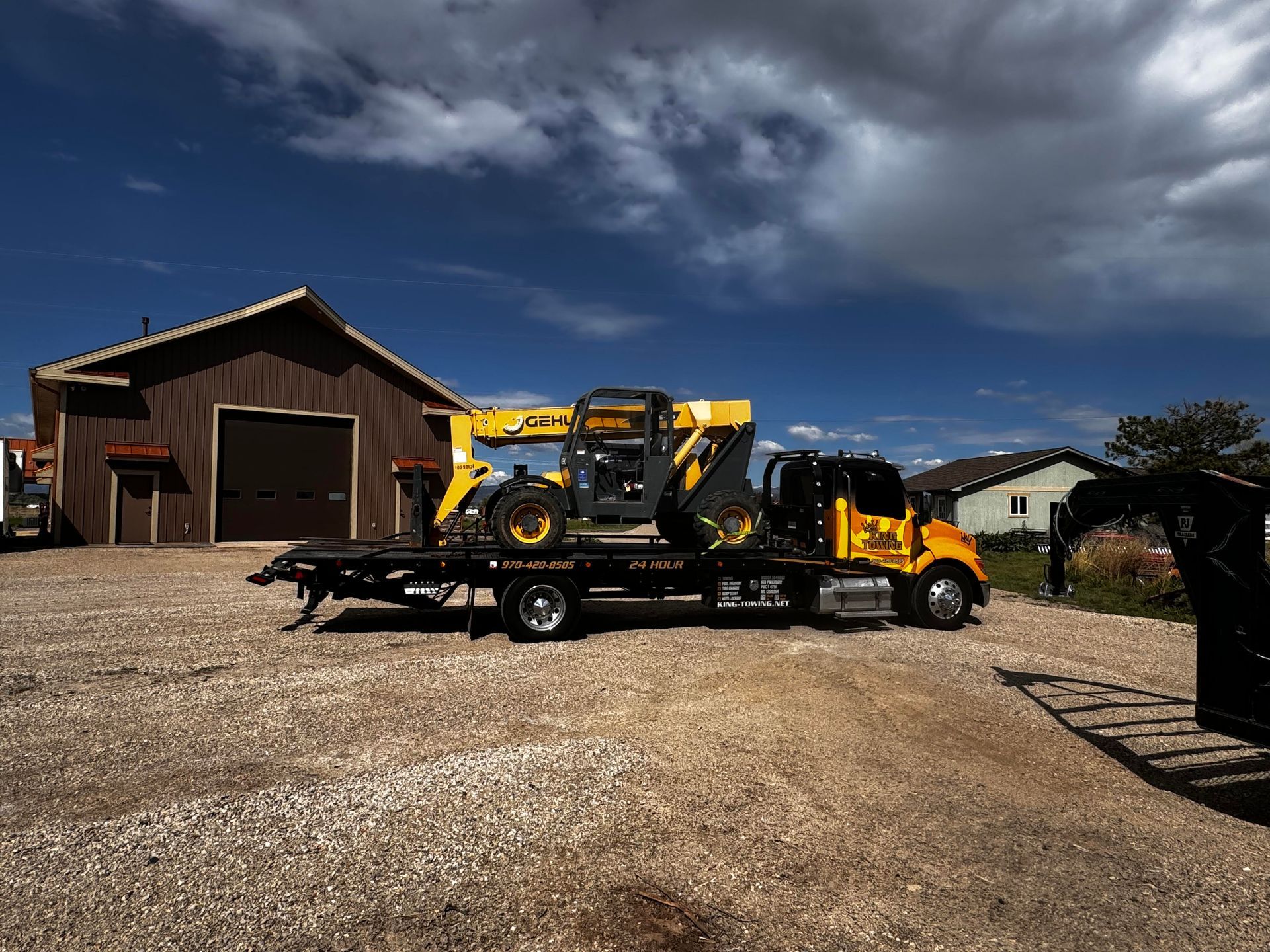 Yellow tow truck carrying a skid steer on a trailer in front of a barn under a cloudy sky