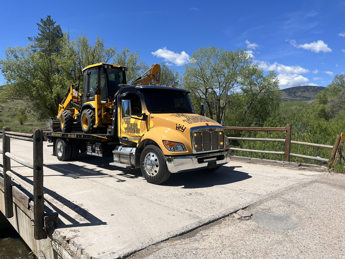 Yellow backhoe loader on a bridge over a creek, with trees and mountains in the background