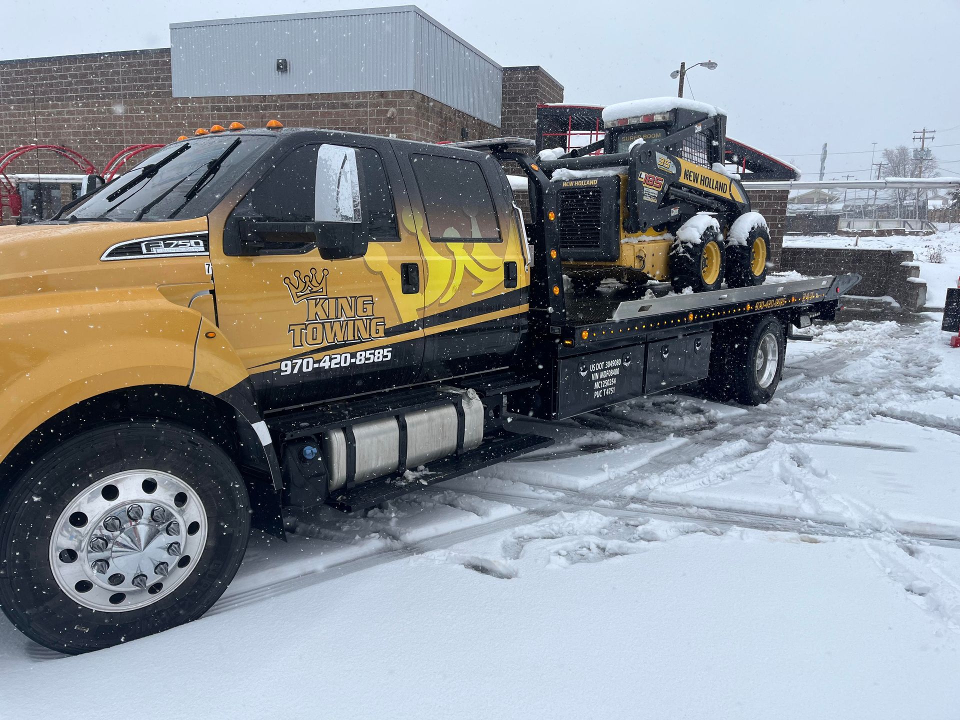 Yellow tow truck parked in snowy lot with car on flatbed under falling snow
