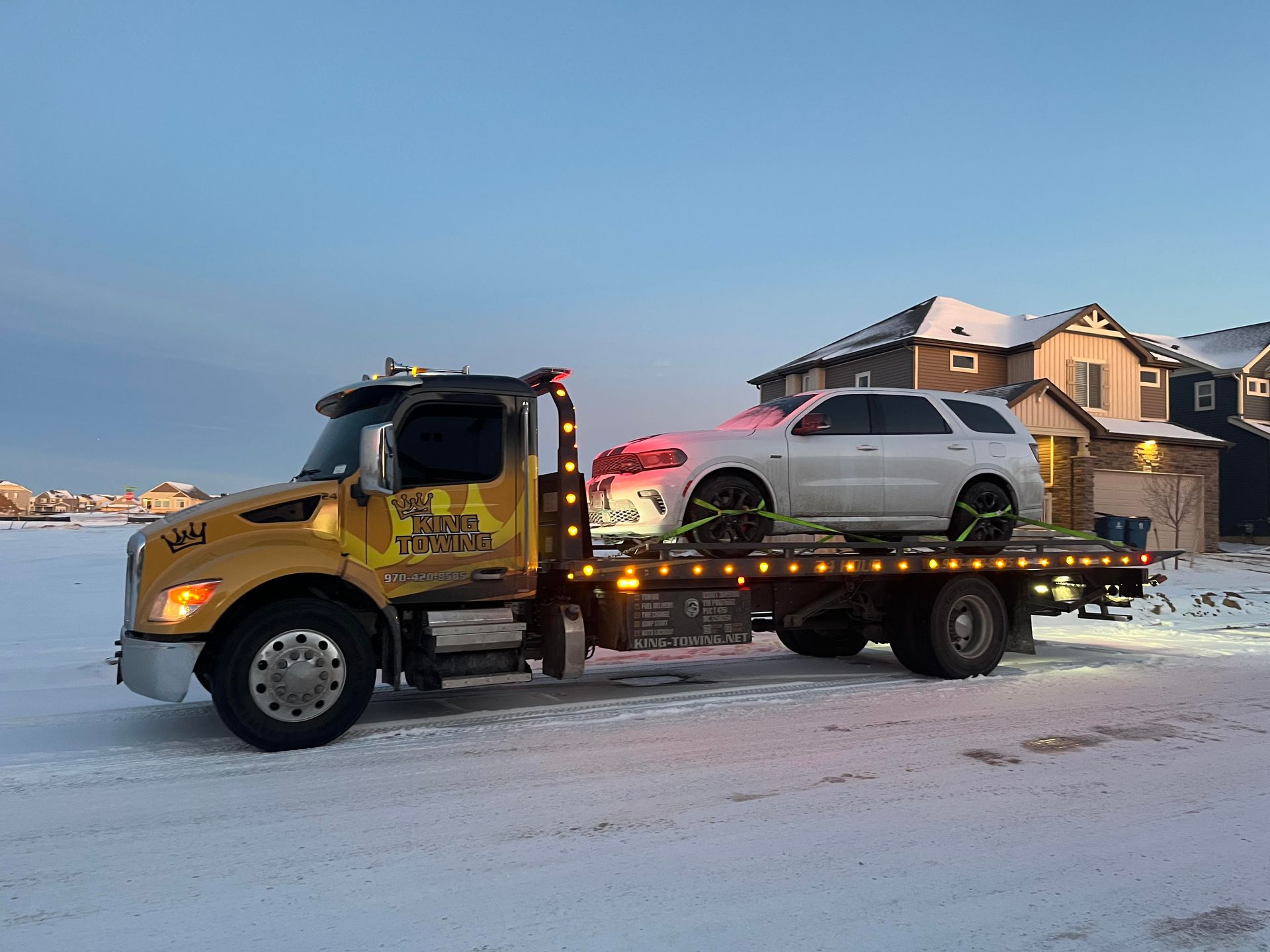 Tow truck hauling a silver SUV on a snowy street beside a lit house at dusk