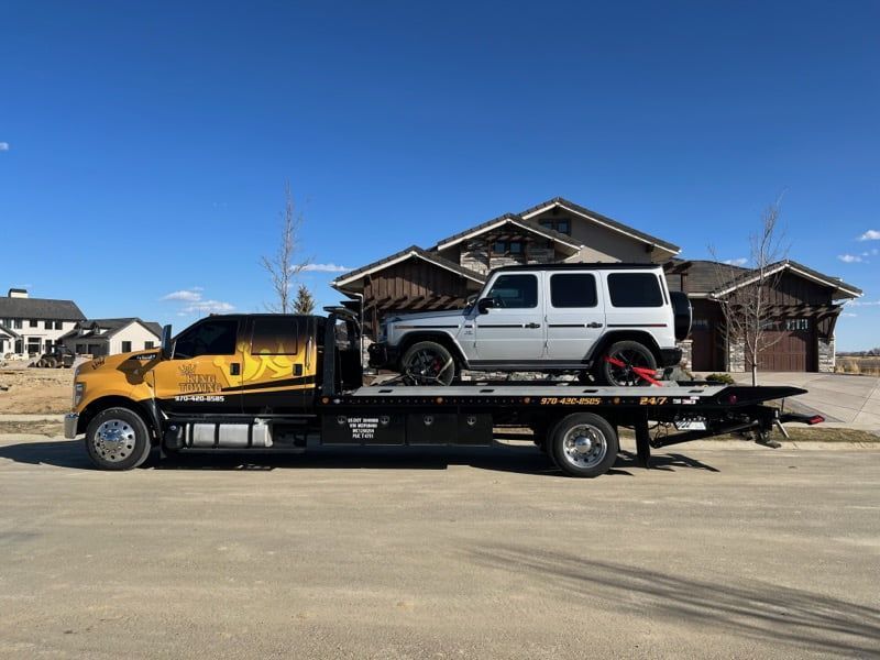Flatbed tow truck carrying a silver SUV on a road beside houses under a clear blue sky