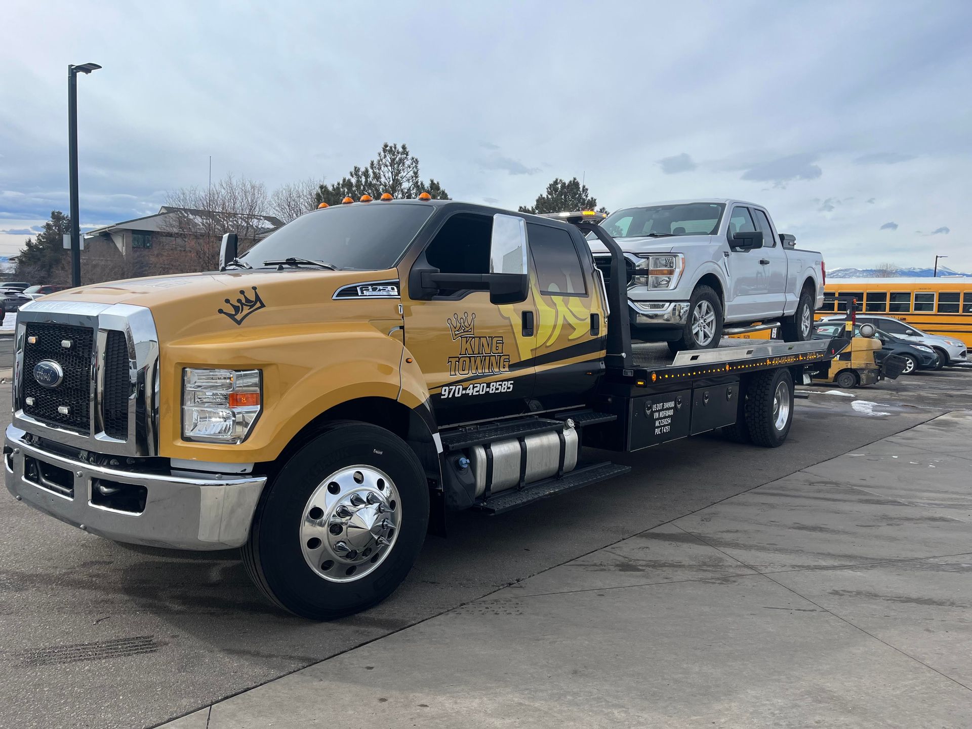 Yellow flatbed tow truck carrying a white SUV on a parking lot under a cloudy sky