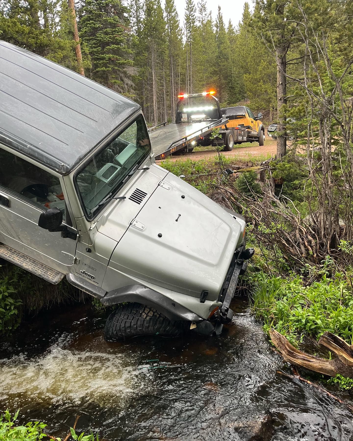 White SUV nose-down in a creek beside a forest road, with a tow truck in the background.