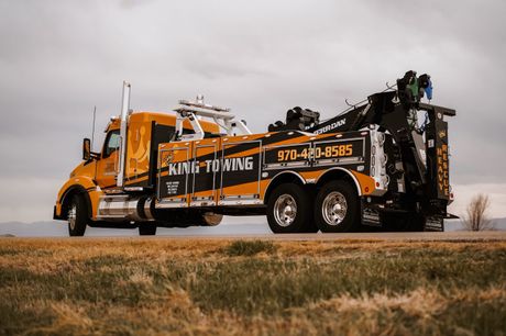 Orange tow truck with a crane arm parked on a road beside a grassy field under an overcast sky