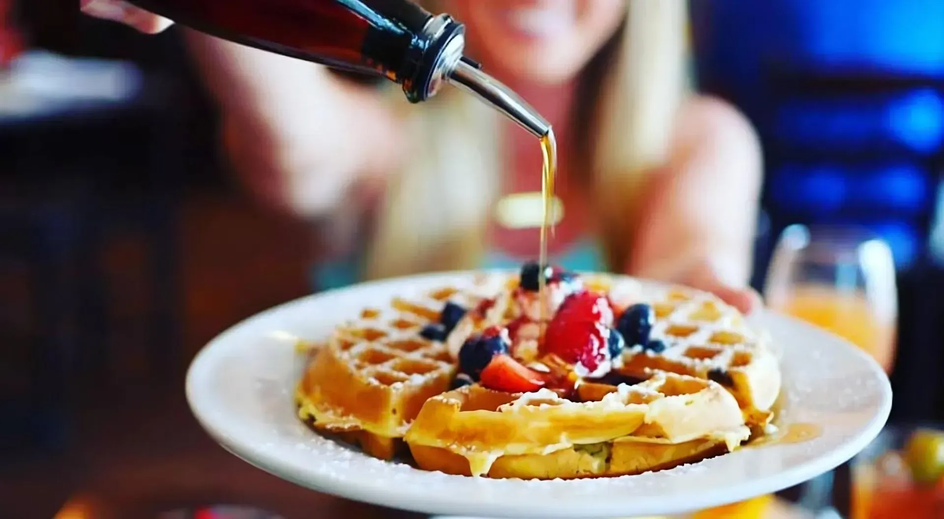 A woman is pouring syrup on a waffle with strawberries and blueberries.