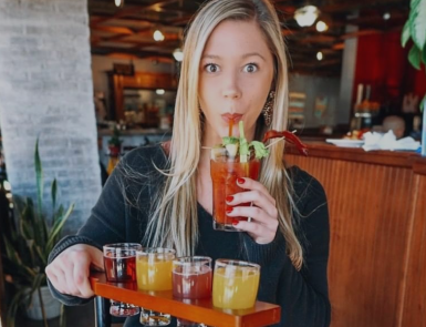 Woman in a restaurant holding a tray of colorful drinks, sipping a red cocktail with a surprised expression
