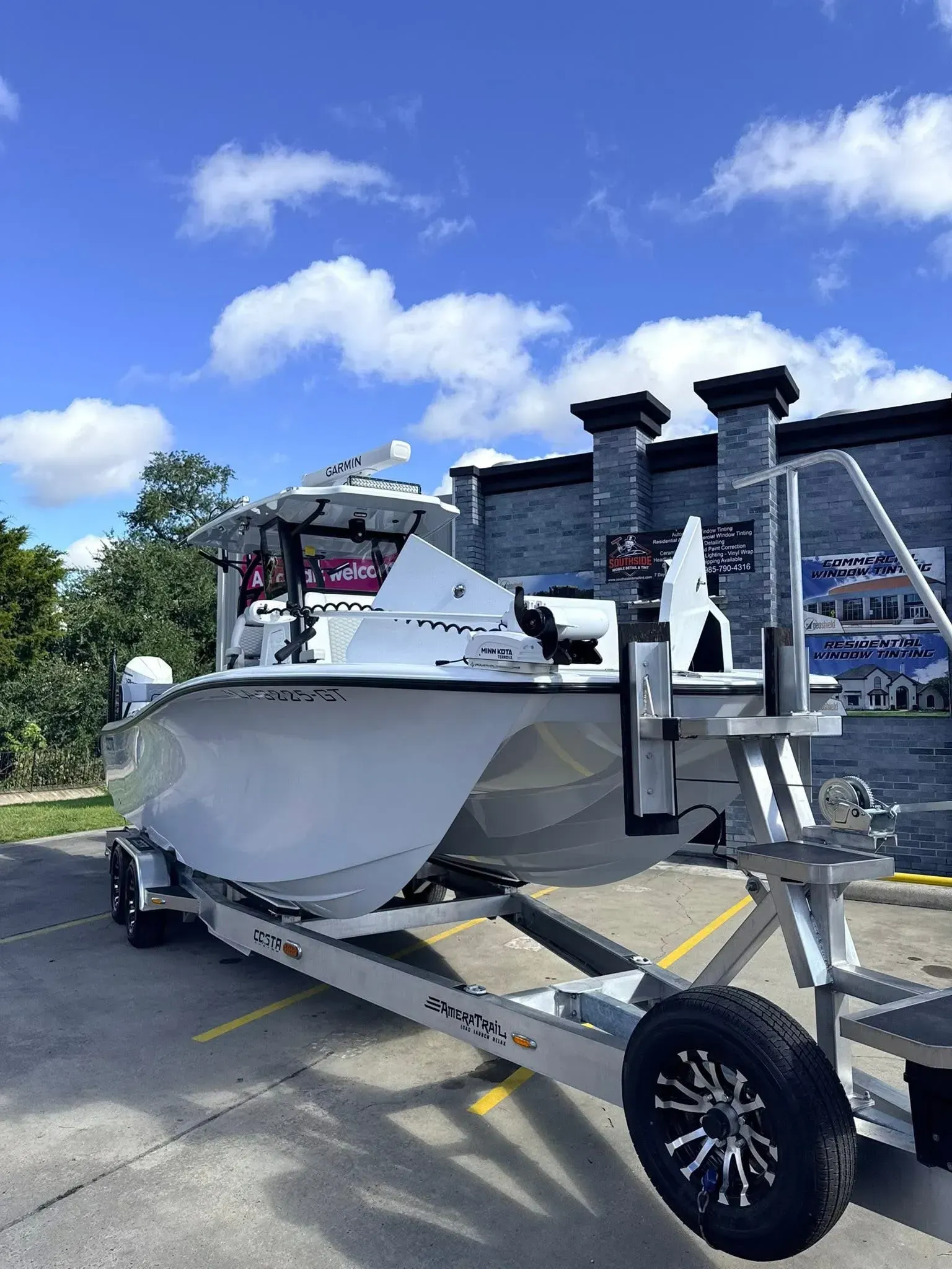 White boat on a trailer, parked on pavement against a backdrop of a building and a blue sky with clouds.