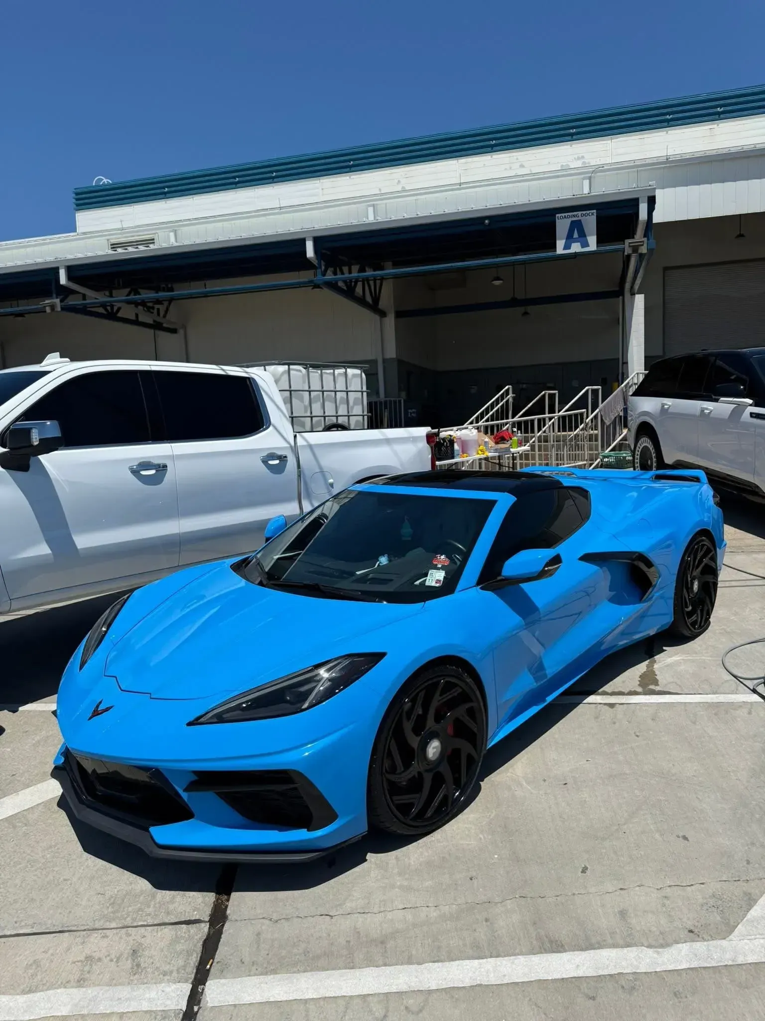 Blue Chevrolet Corvette convertible parked in front of a building with a white truck visible.