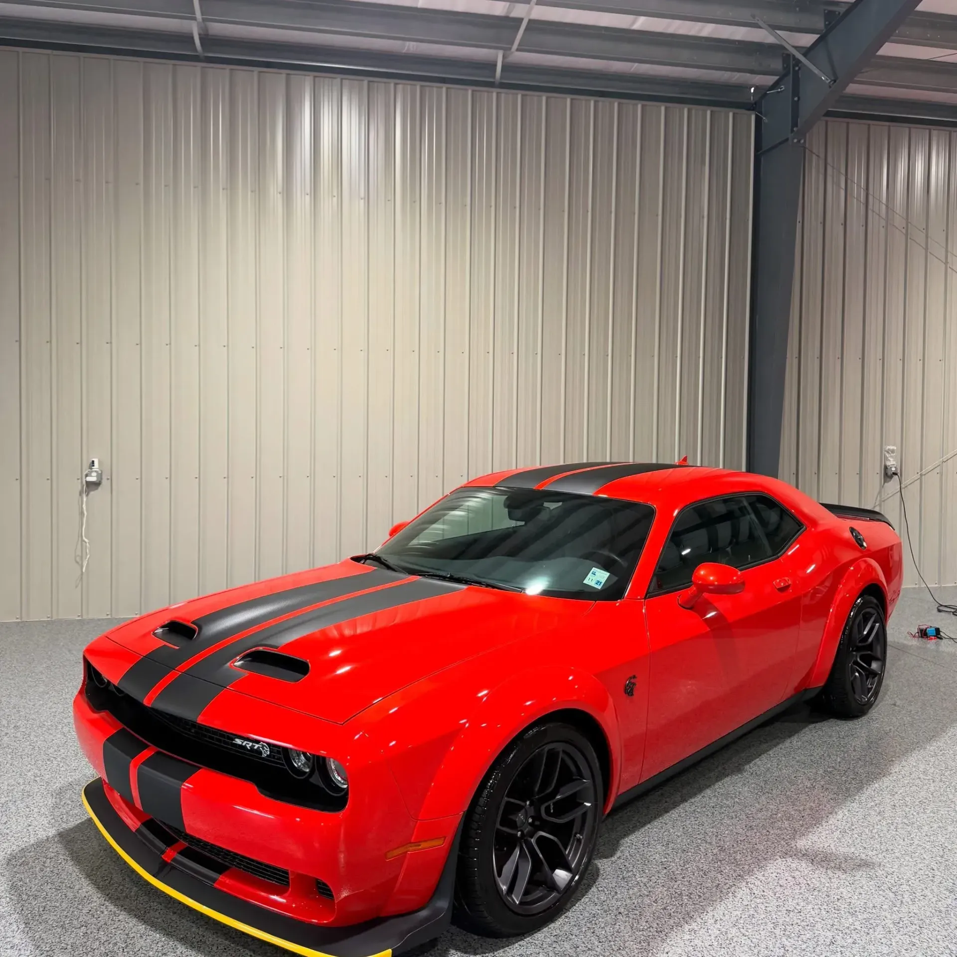 Red Dodge Challenger with black stripes parked inside a garage with tan walls and a gray floor.
