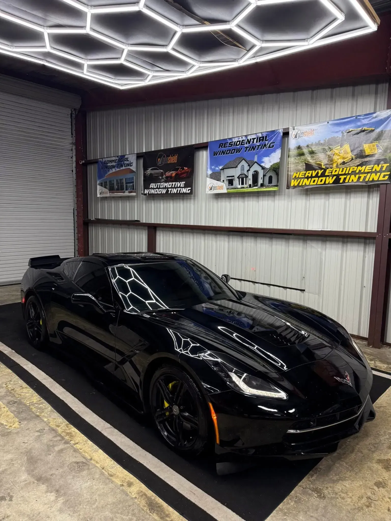 Black sports car in a well-lit garage, featuring hexagonal lights and promotional banners on the wall.