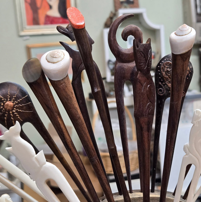 Close-up of a collection of ornate wooden hair sticks with carved animal and shell designs in a shallow container.