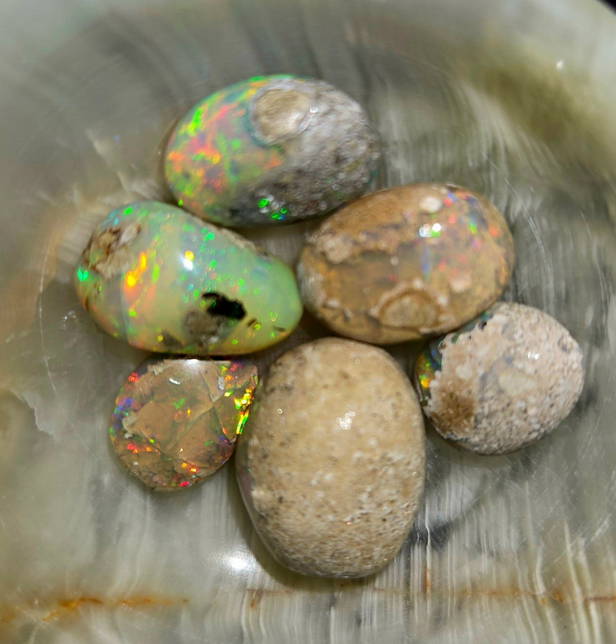 Six rough opals in a marble dish, displaying flashes of iridescent color against their earthy exteriors.