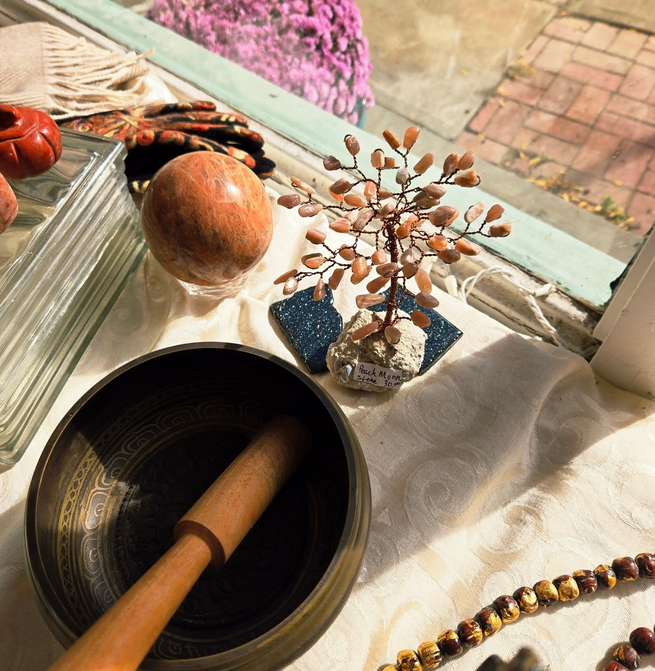 Tibetan singing bowl with mallet, a stone tree sculpture, and a rock ball on a white cloth near a window.