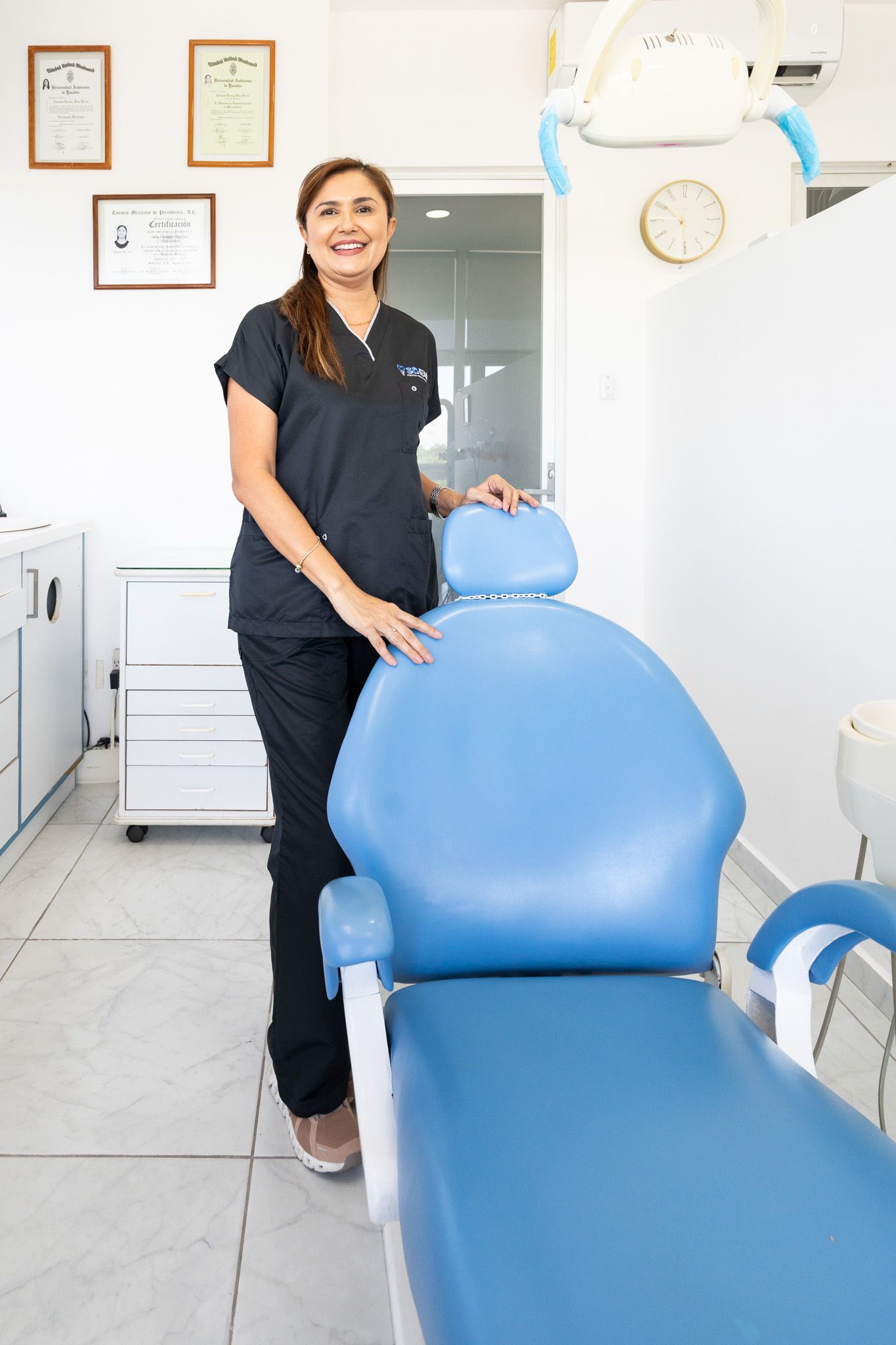 Dentista de pie junto a un sillón dental azul en una luminosa sala de consulta.