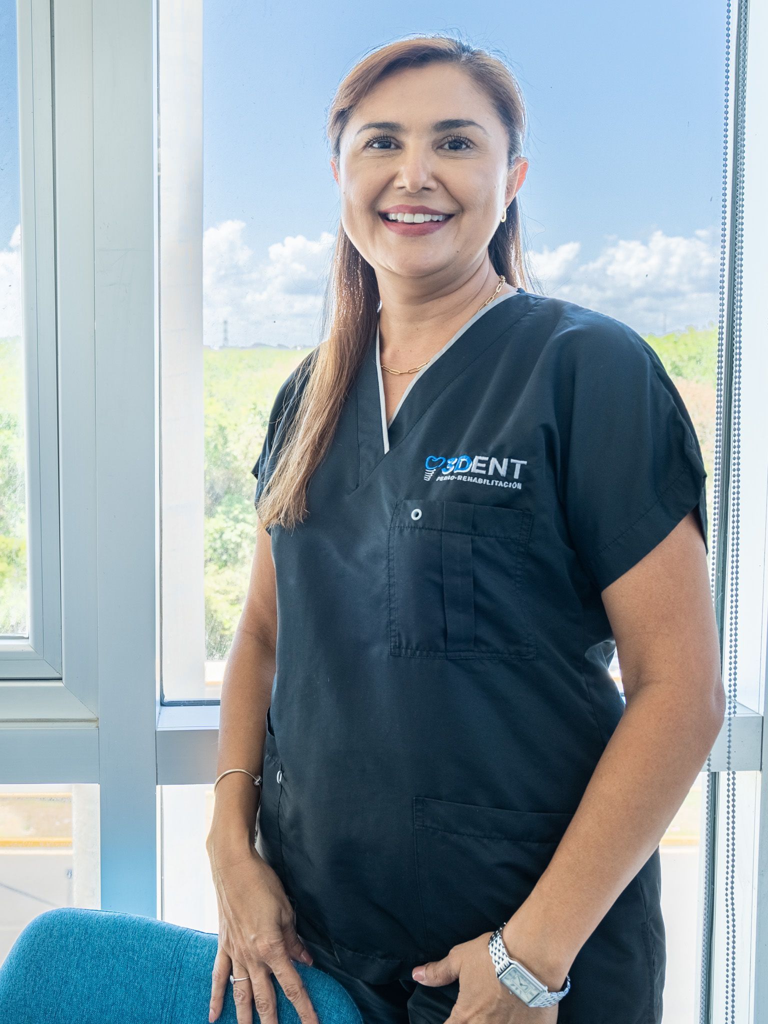 Mujer con uniforme médico negro sonriendo junto a una ventana luminosa, de pie al lado de una silla azul.