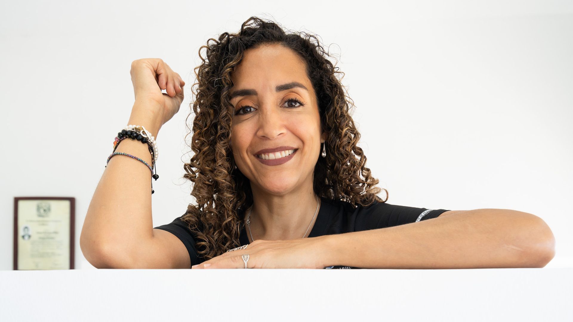Persona sonriente con cabello rizado flexionando un brazo sobre un fondo blanco, con un certificado enmarcado a la izquierda.