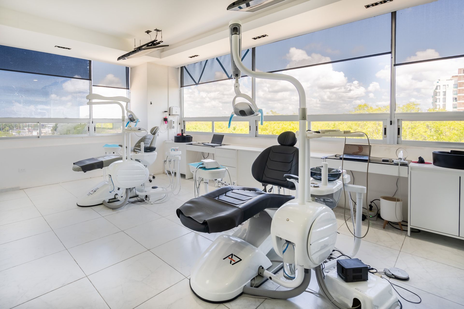 Clínica dental moderna y luminosa con dos sillones de exploración blancos y grandes ventanales.