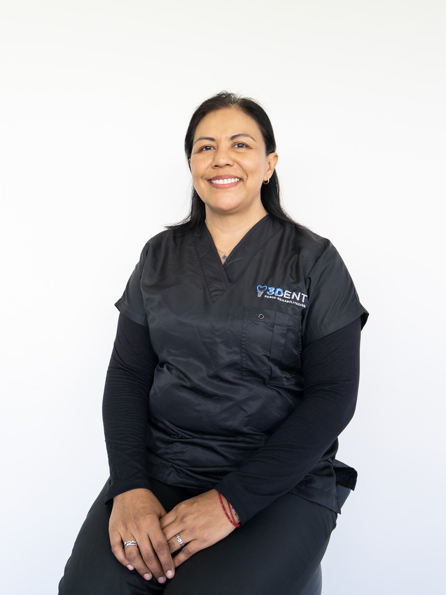 Mujer sonriente con uniforme médico negro sentada sobre un fondo blanco.
