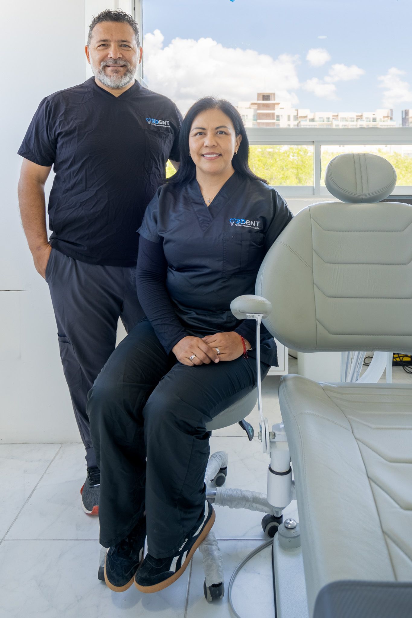 Dos personas con uniformes negros posando en una luminosa consulta médica o dental.