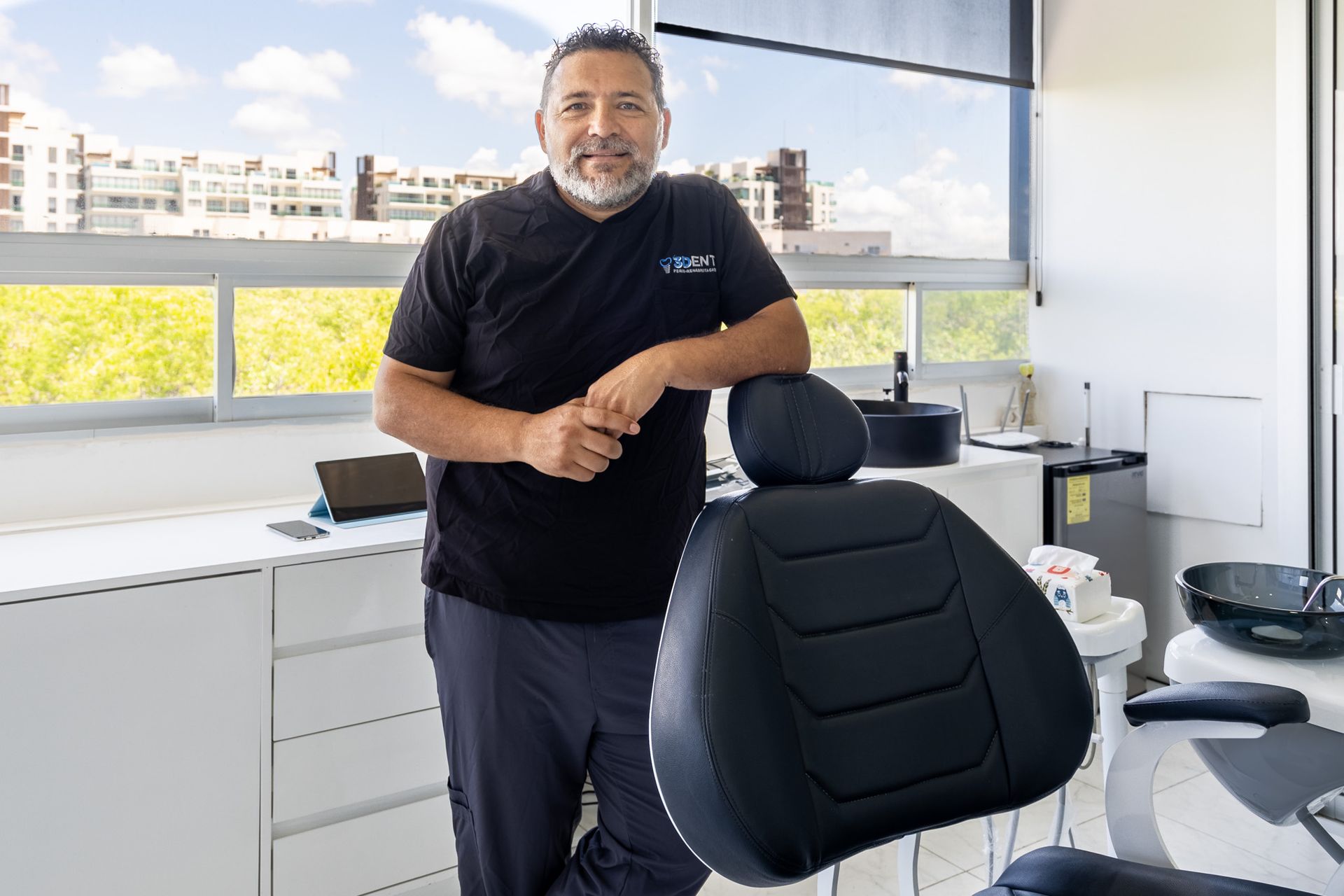 Un hombre de pie en una oficina moderna junto a una silla negra, con grandes ventanales y vistas a la ciudad detrás de él.