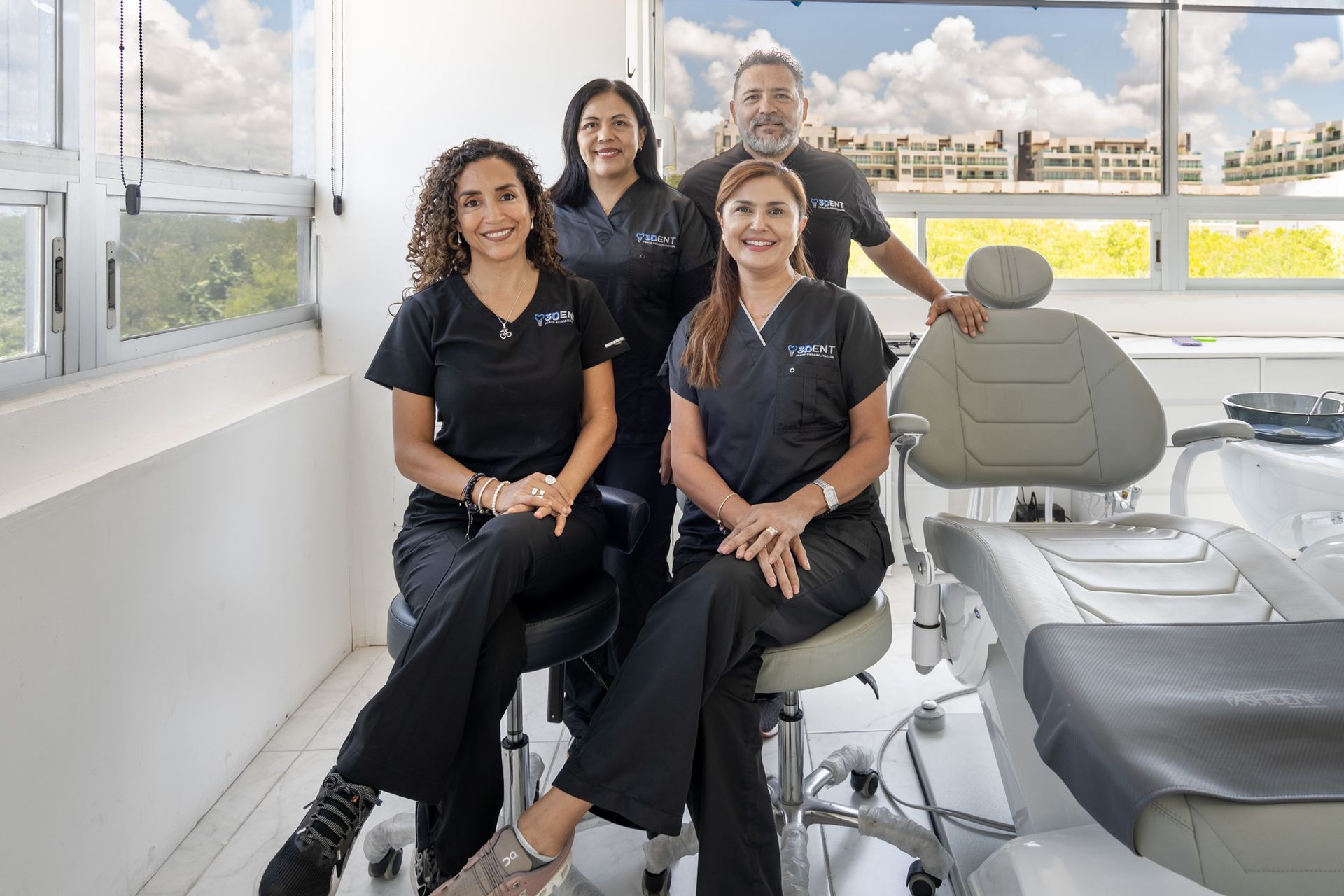 Cuatro profesionales dentales sonríen en una clínica moderna y luminosa con sillones dentales y grandes ventanales.