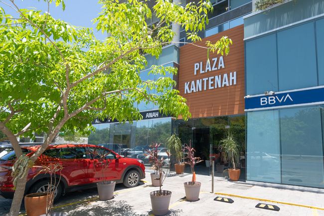 Fachada de un local en Plaza Kantenah con un SUV rojo, plantas en macetas y un letrero de BBVA en una calle soleada.