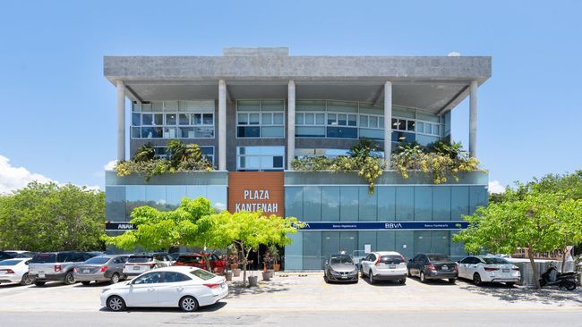 Edificio moderno con fachada de cristal azul, árboles y coches aparcados bajo un cielo despejado.