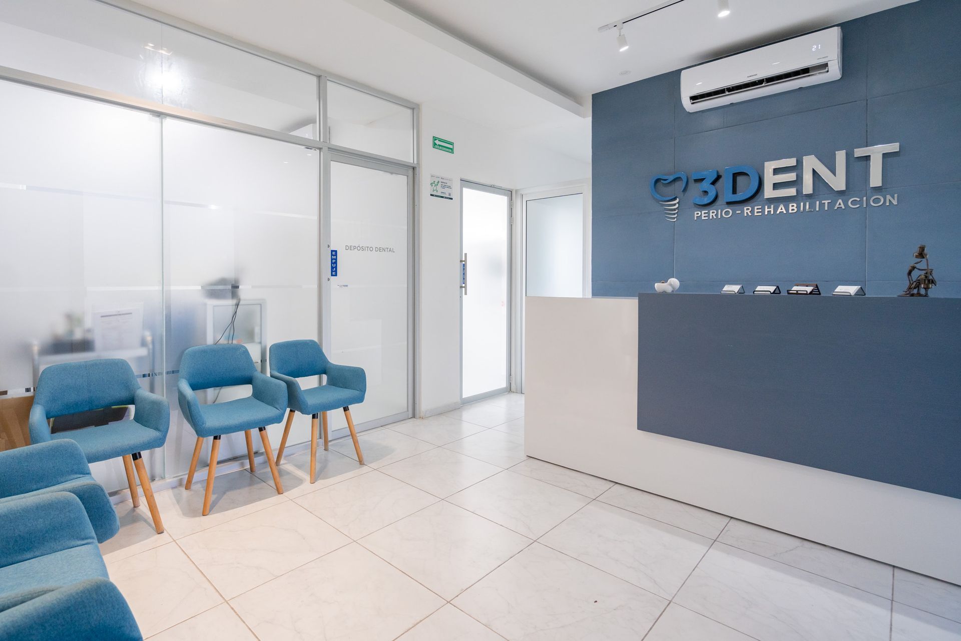 Recepción moderna de una clínica dental con sillas azules, sala de espera con paredes de cristal y suelo de baldosas blancas.