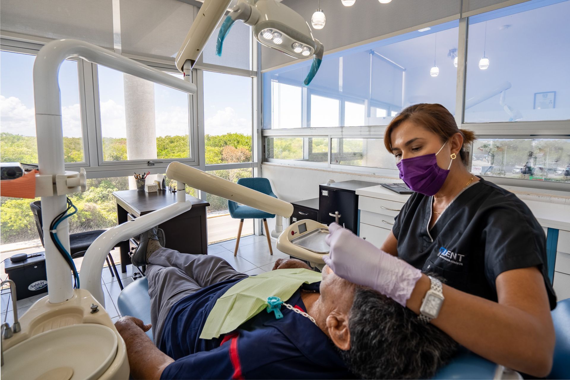 Dentista atendiendo a un paciente en una clínica luminosa, con mascarilla y guantes, con instrumental dental encima.