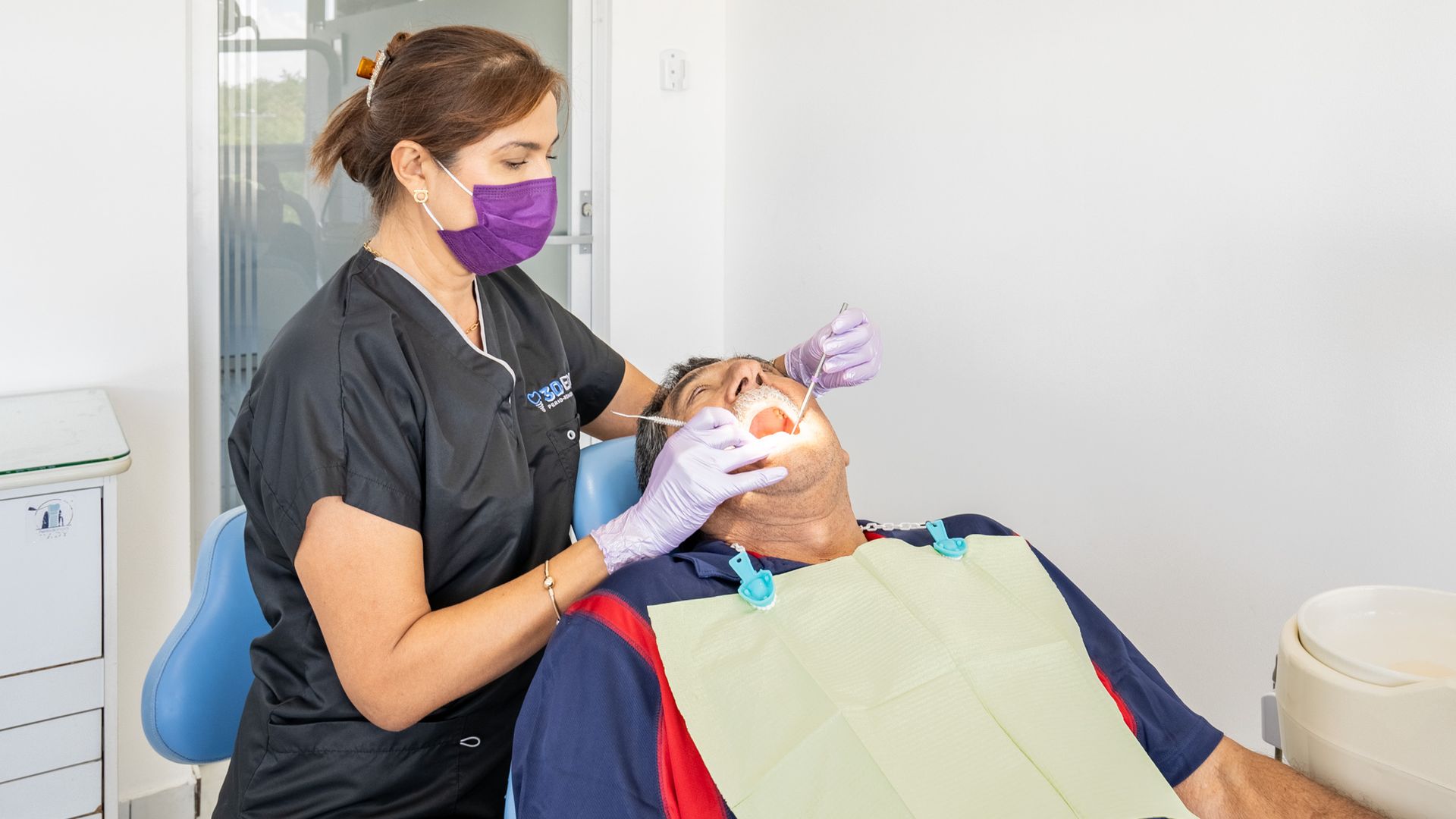 Dentista con mascarilla y guantes morados atendiendo a un paciente recostado en una clínica.
