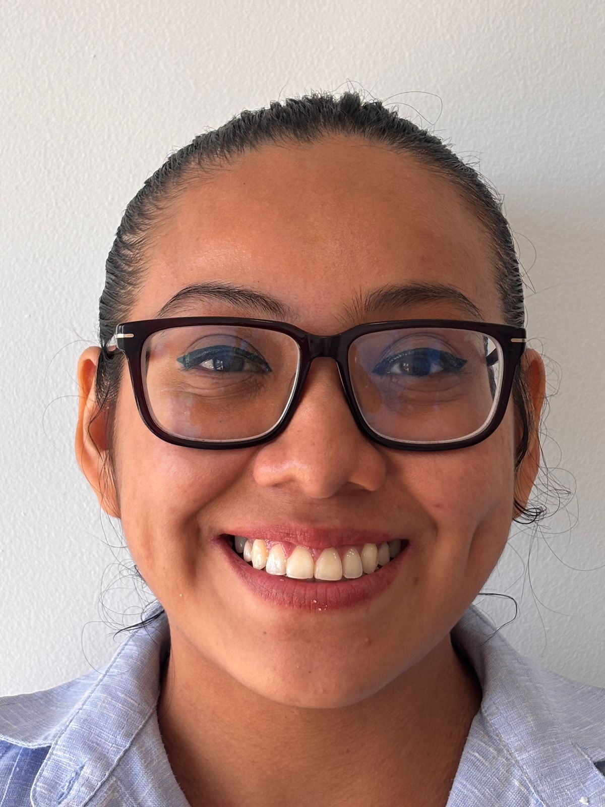 Persona sonriente con gafas negras y camisa azul claro contra una pared blanca lisa.