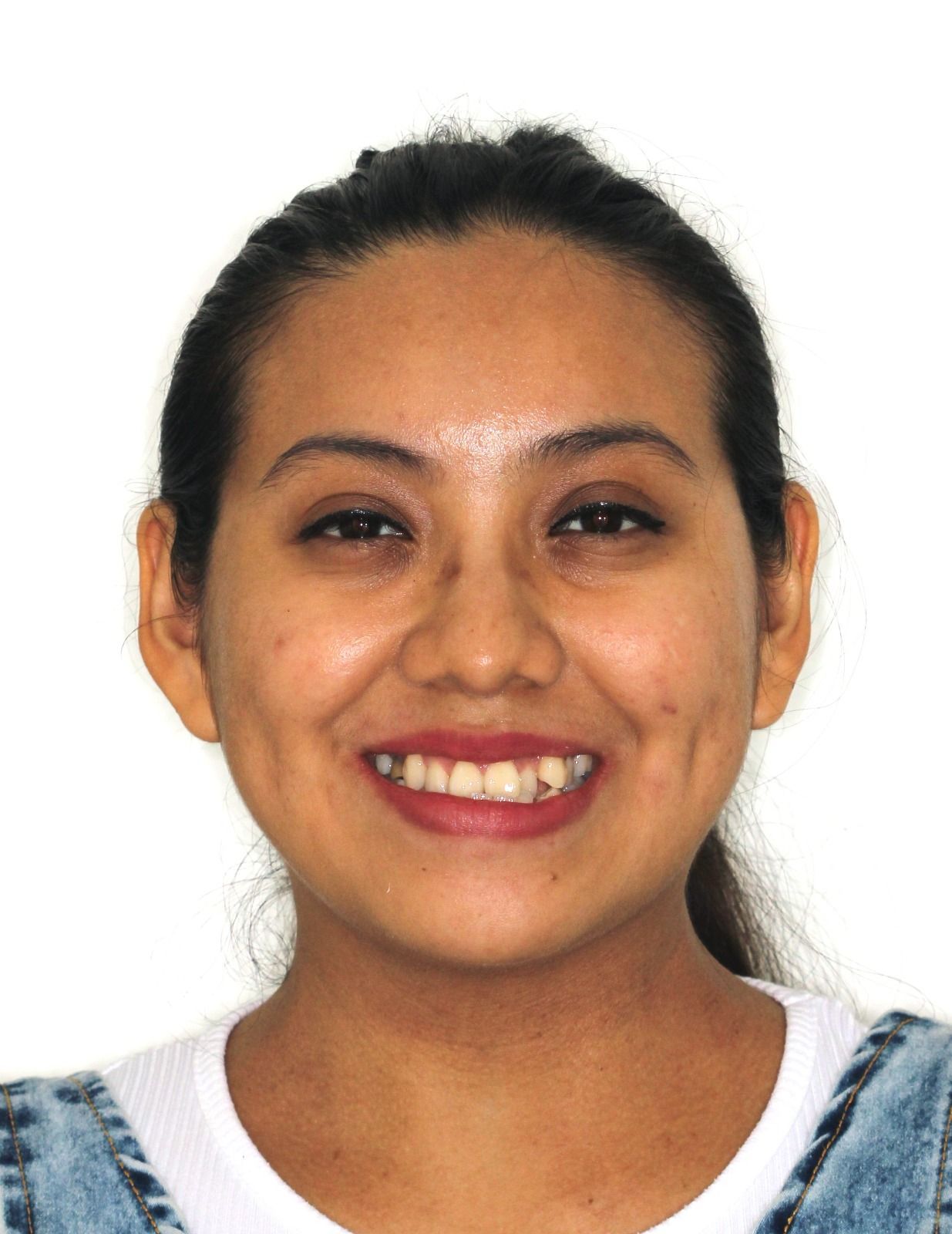 Persona sonriente de cabello oscuro sobre fondo blanco, vestida con una camiseta blanca y un mono vaquero.