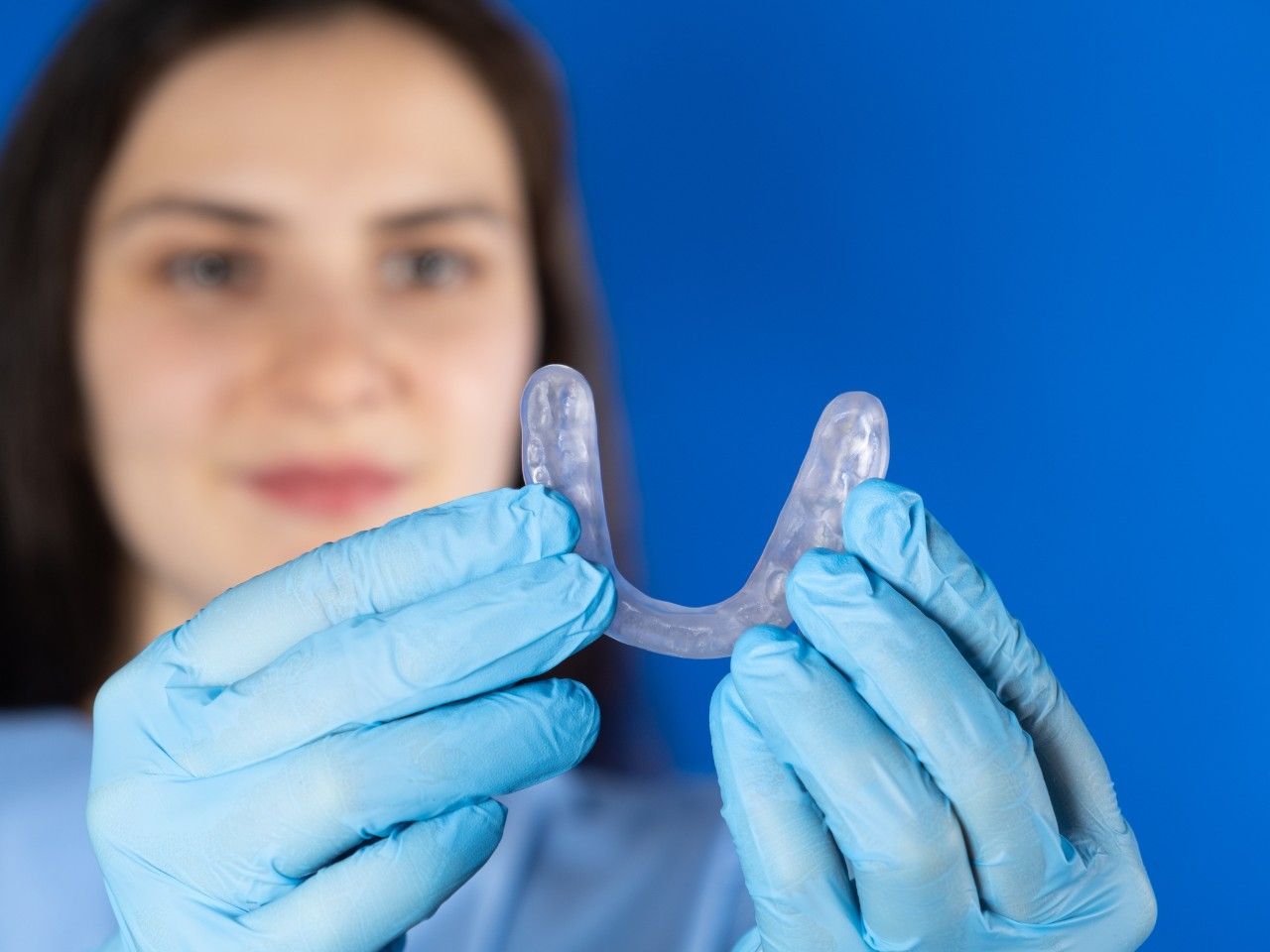 Manos enguantadas sosteniendo un retenedor dental transparente frente al rostro borroso de una persona.