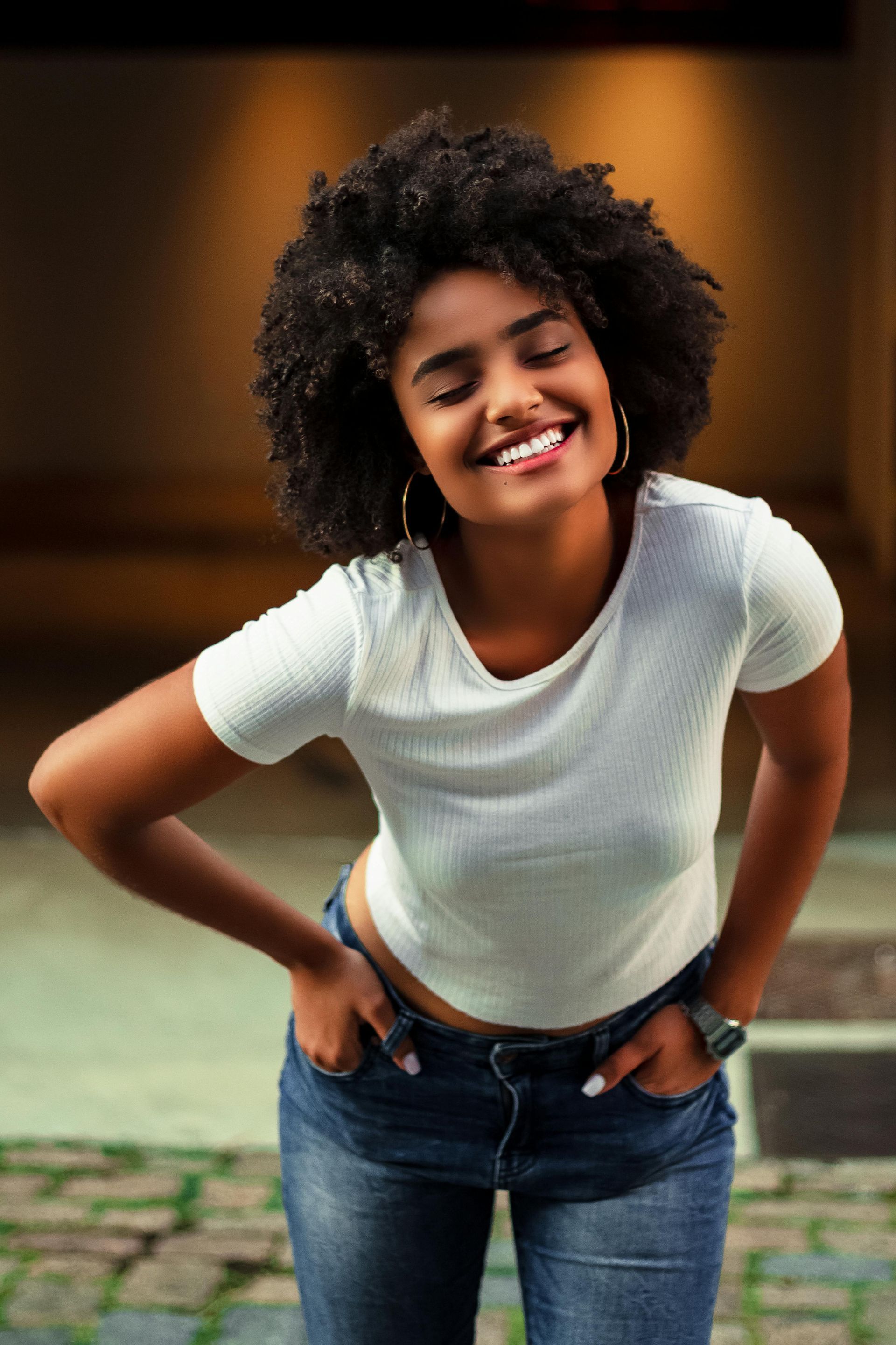 Persona sonriente con camisa blanca y vaqueros, inclinada hacia adelante con las manos en las caderas, al aire libre.