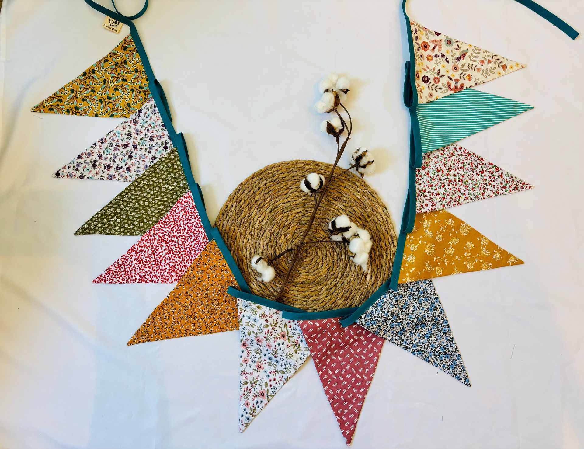 Kleurrijke driehoekige slingers rond een goudkleurig geweven ornament met witte bloemen op een witte achtergrond.