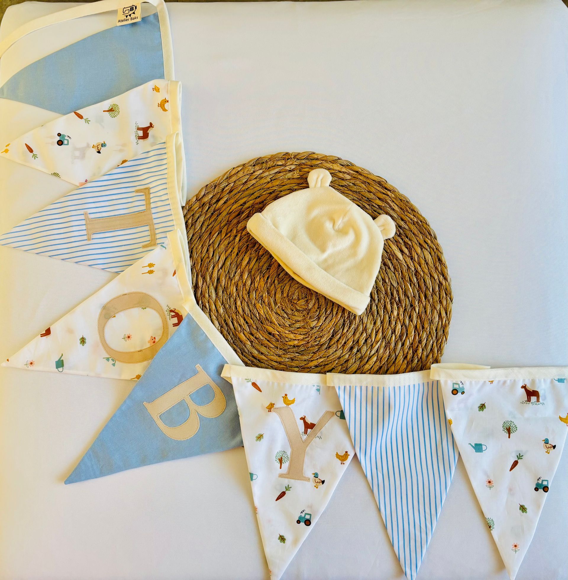 Pastelkleurige vlaggetjes met babymotieven naast een geweven placemat en een witte decoratieve roos op een lichte achtergrond.