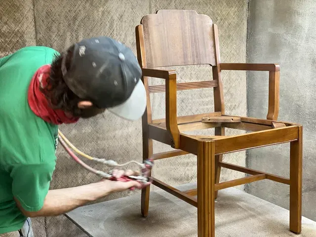 A man in a green shirt is spray painting a wooden chair.