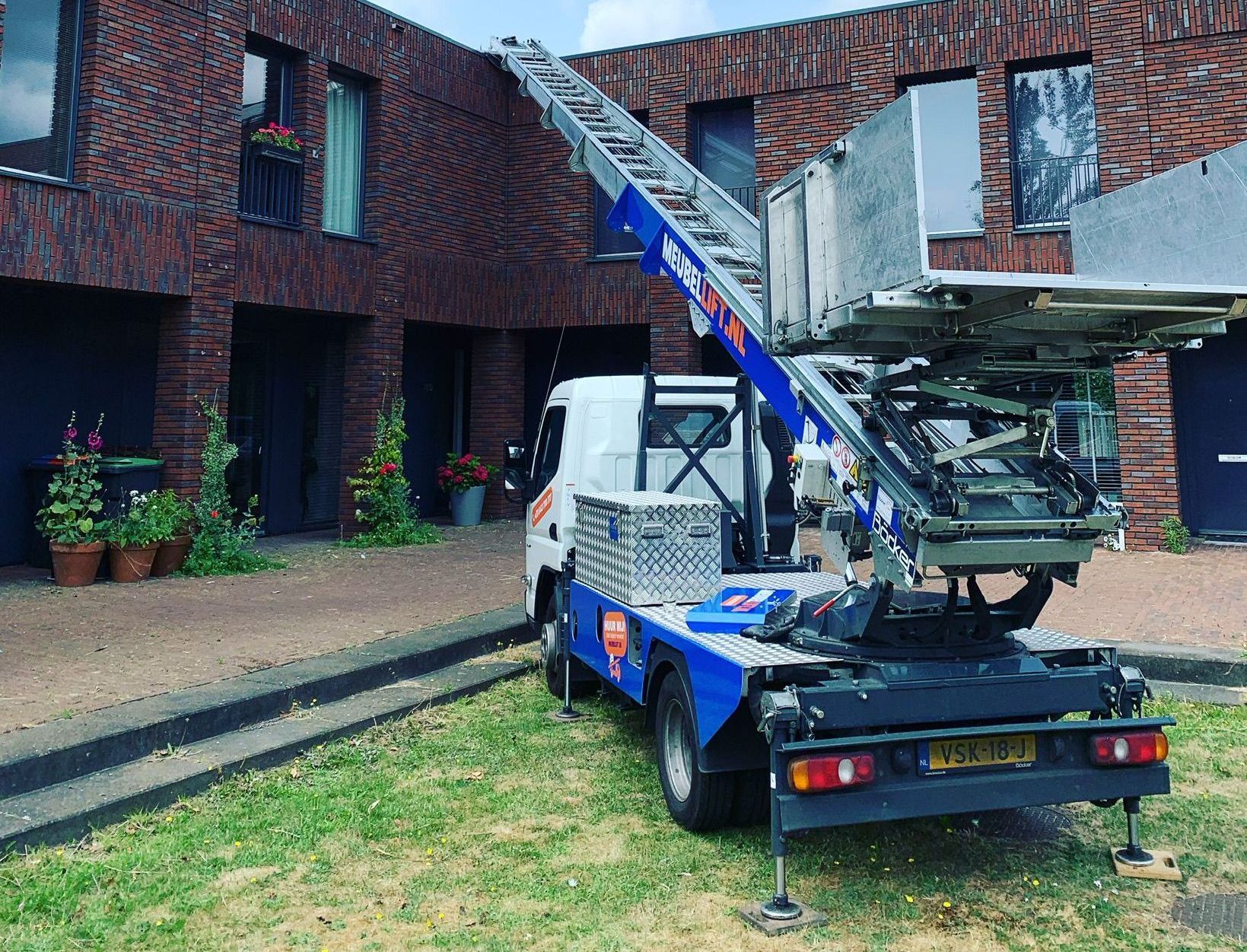 Een blauw-witte vrachtwagen met een kraan aan de achterkant staat geparkeerd voor een bakstenen gebouw.